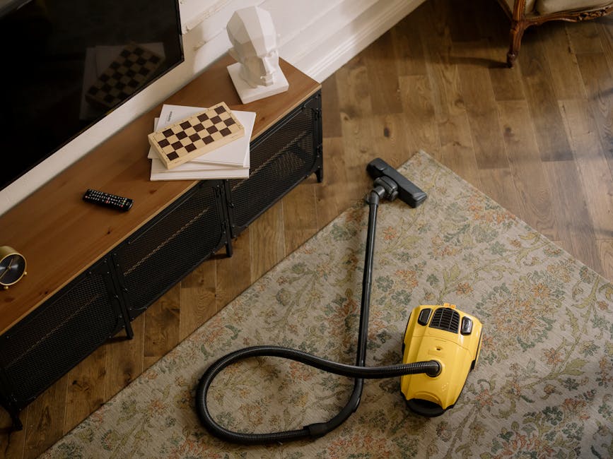 A man with dark, curly hair and wearing a burnt orange long-sleeve shirt and olive green pants is performing surface cleaning on a light grey fabric sofa in a living room. He is bent over, carefully p