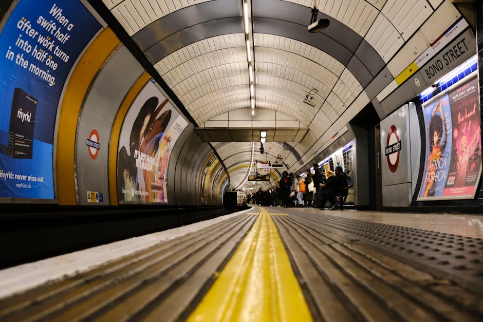 Wide-angle view of Colliers Wood Tube Station platform in SW19, showing the curved tiled ceiling illuminated by linear fluorescent lights. The platform has a yellow safety line along its edge and a textured tactile paving strip for visually impaired passengers. Advertisements are displayed on the platform walls, including a blue campaign poster and a colorful fashion ad. Several passengers are seated and standing near the station's edge, waiting for the train. The cleanliness of the platform appears well-maintained with no visible litter or dirt, reflecting routine surface cleaning and sanitisation by professional cleaning services such as Carpet Cleaners SW19. The image highlights the subway environment's hygiene and safety features, with a focus on the tiled flooring and detailed platform surface.