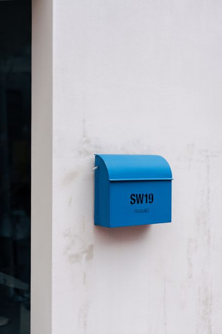 A blue mailbox mounted on a white exterior wall near Colliers Wood Tube Station, marked with the postal code SW19 and the word 'FRAGILE'. The wall shows slight signs of dirt and weathering, and the mailbox has a smooth, curved top with a small slot for mail. The lighting is natural, suggesting an outdoor setting, and the scene is clean and well-maintained, reflecting attention to hygiene and upkeep often associated with professional cleaning and surface sanitisation. This image is relevant to the focus of surface cleaning and maintenance for residential and commercial properties, as highlighted in the Colliers Wood Tube Station carpet cleaning guide SW19 by Carpet Cleaners SW19.