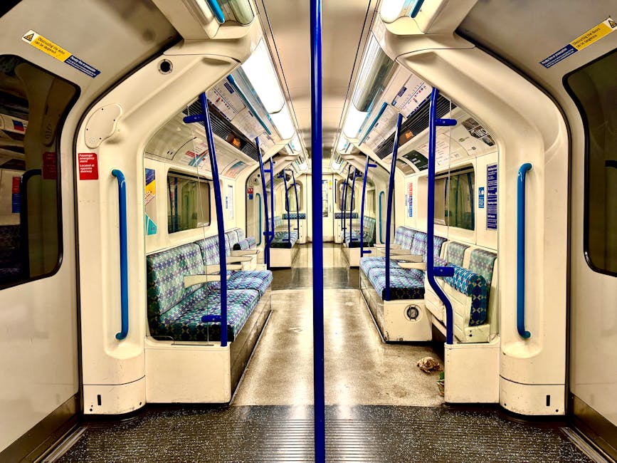 Wide-angle view of Colliers Wood Tube Station platform in SW19, showing the curved tiled ceiling illuminated by linear fluorescent lights. The platform has a yellow safety line along its edge and a textured tactile paving strip for visually impaired passengers. Advertisements are displayed on the platform walls, including a blue campaign poster and a colorful fashion ad. Several passengers are seated and standing near the station's edge, waiting for the train. The cleanliness of the platform appears well-maintained with no visible litter or dirt, reflecting routine surface cleaning and sanitisation by professional cleaning services such as Carpet Cleaners SW19. The image highlights the subway environment's hygiene and safety features, with a focus on the tiled flooring and detailed platform surface.