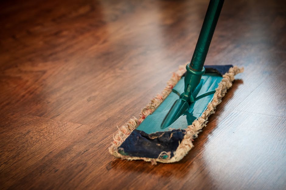 A cleaning mop with a green handle and a blue wringing mechanism is positioned on a polished wooden floor. The mop pad, made of a fabric material with frayed edges, appears used but clean. The surface